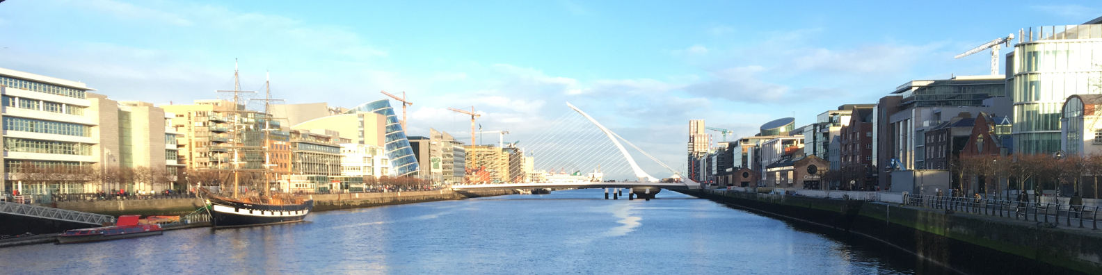 A panorama photo of the river Liffey in Dublin taken from the Sean O'Casey Bridge. Central in the photo is the iconic Samuel Beckett Bridge, which is shaped like the harp that is one of Ireland's national symbols. Also in the photo is the tall ship, Jeanie Johnston.